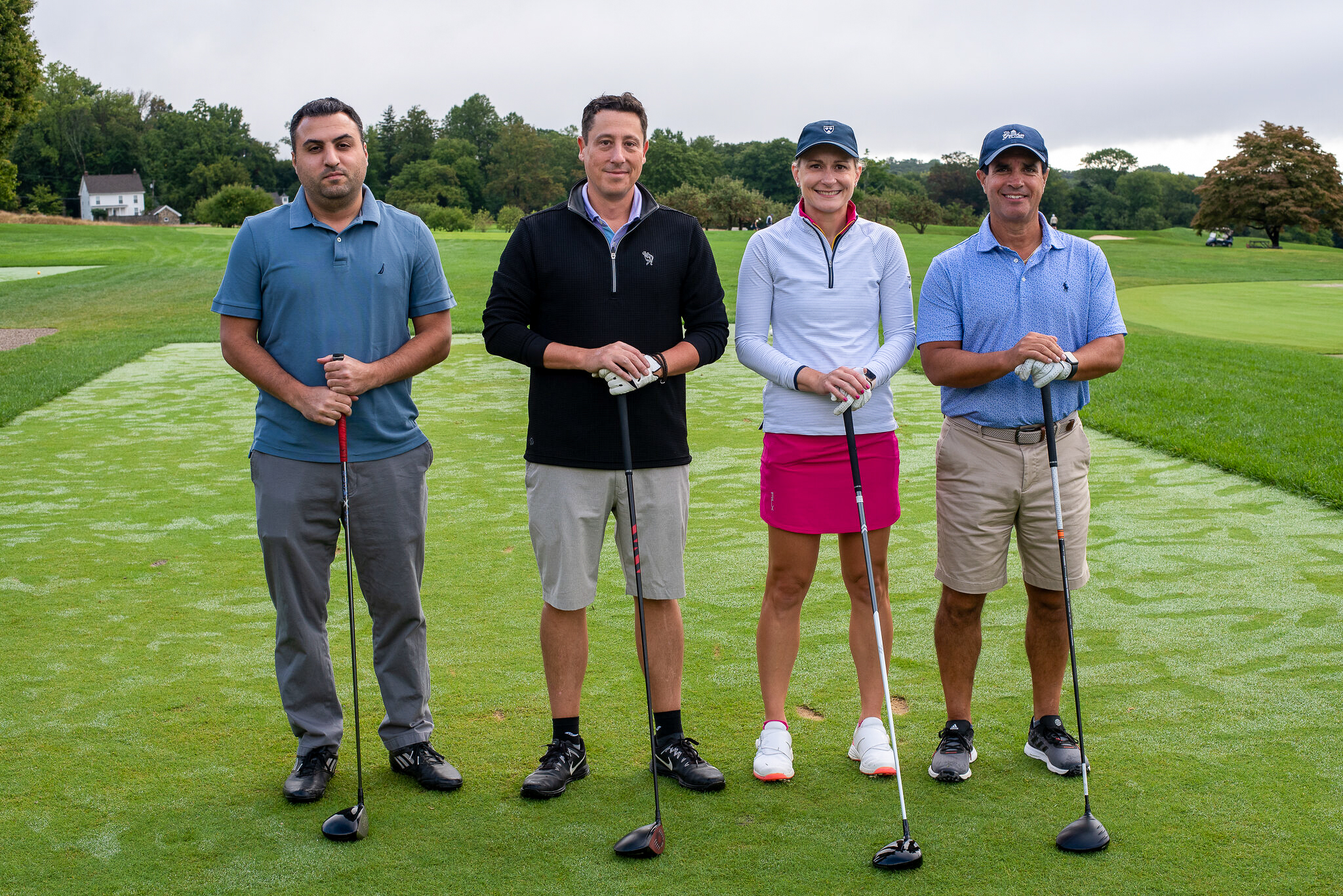 Four people stand with golf clubs, facing the camera, in front of a golf course