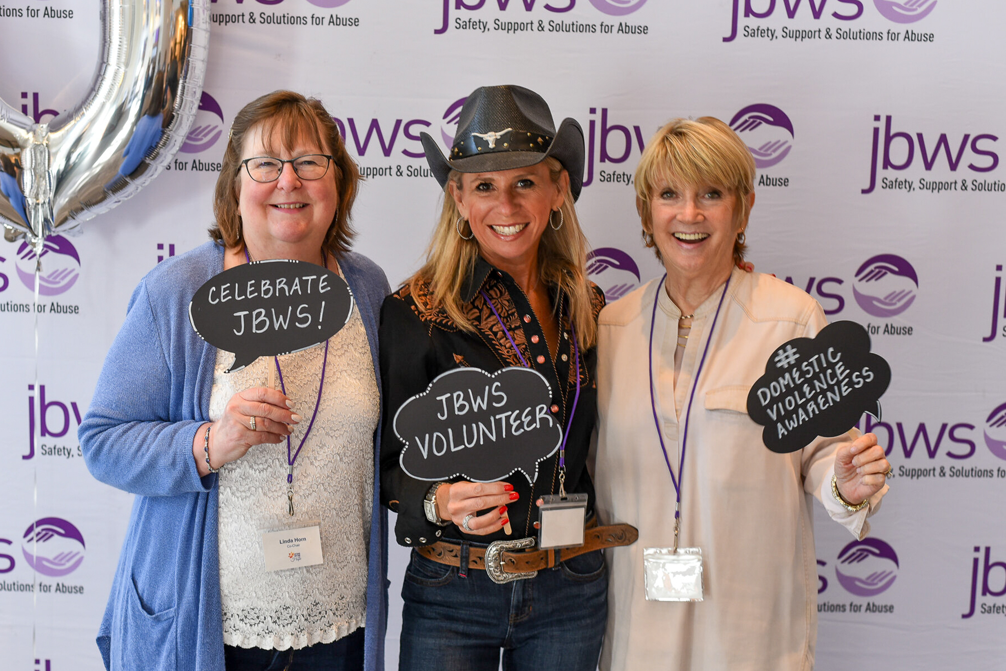 Three women holding signs that say "JBWS volunteer" stand before a step and repeat