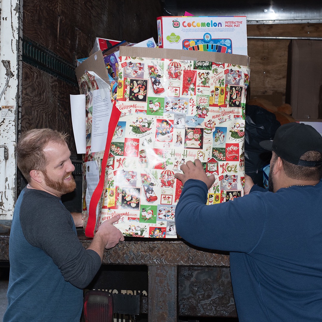 Two men are lifting a huge box, wrapped in Christmas wrapping paper, from a box truck